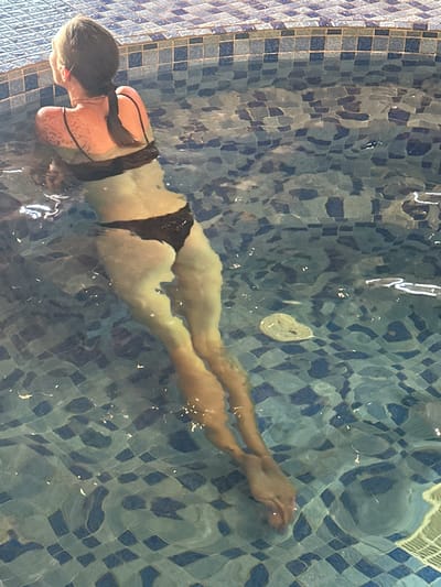 This media file features an overhead view of a person submerged in a swimming pool, actively swimming or floating. The subject is a female, visible from head to toe, wearing a dark two-piece swimsuit. Her body is elongated in a prone position, with arms and legs extended.
The swimming pool's interior is decorated with blue and white/light blue square tiles, forming a checkered pattern on both the bottom and the visible side wall. The water is clear, allowing the pool's features and the swimmer to be seen distinctly, but its surface is dynamic. Significant ripples and numerous bright reflections are visible on the water's surface, creating wavy distortions of the tiled pattern and the swimmer's body, along with streaks of light.
A small, light-colored, amorphous oval object is seen floating near the surface in the upper central area of the pool. The overall lighting appears bright and consistent, characteristic of either a well-lit indoor pool or a covered outdoor setting. There are no discernible emotional expressions, specific time of day or weather conditions visible, nor any visible text in the image. The provided location context is Mu Cang Chai, Vietnam, but no visual cues within the image confirm this specific geographical location.