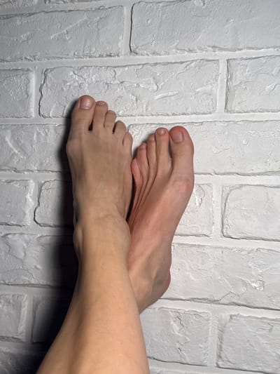 The image displays two bare feet against a white brick wall. The feet are positioned side-by-side, with one foot slightly overlapping the other. The toes are visible, and the skin tone suggests a fair complexion. The brick wall shows texture and grout lines, indicating an indoor or outdoor setting. There is no information in the image to suggest the location in the city of Lipetsk, Russia.