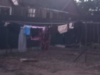 The image depicts an outdoor scene in what appears to be a rural setting, likely in Кенжекөл, Kazakhstan, given the provided location context. The primary subjects are laundry items hanging on a clothesline strung across the foreground. These include several garments, with some appearing to be shirts or dresses, in various colors such as light gray, blue, pink, and a beige or off-white fabric. The laundry is draped over the line, suggesting it is either drying or has recently been washed.
Behind the clothesline, the background reveals a dimly lit, rustic dwelling, possibly a house or barn, with visible windows that are either dark or reflect faint light. The structure appears weathered and somewhat indistinct due to the low light conditions. A wooden fence is partially visible to the right of the frame, further contributing to the rural ambiance. The ground in the foreground is covered in dirt and scattered debris, indicating a natural or unpaved area.
The overall lighting suggests that the photograph was taken during either dawn or dusk, as the natural light is subdued and the sky is not clearly visible, but the presence of some illumination implies it's not pitch black. There is no discernible weather information, though the hanging laundry implies a period without significant rain. No people are visible in the frame. The scene evokes a sense of everyday domestic life in a simple, perhaps modest, living environment. No text is visible in the image.