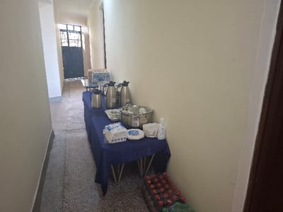 The image shows a narrow hallway with a table set up for a refreshment station. There are four silver thermoses, several silver serving platters, a stainless steel bowl, and a stack of disposable white plates on the table.  There is a small bottle of hand sanitizer, a package of napkins and several packs of bottled water.  The table has a navy blue tablecloth and is positioned against the wall on the right side of the hallway.  There is a closed door at the far end of the hallway and a closed door on the right side of the hallway.  The hallway has beige-colored walls and a tiled floor. The hallway is located in Kisumu, Kenya (latitude: -0.111, longitude: 34.775).