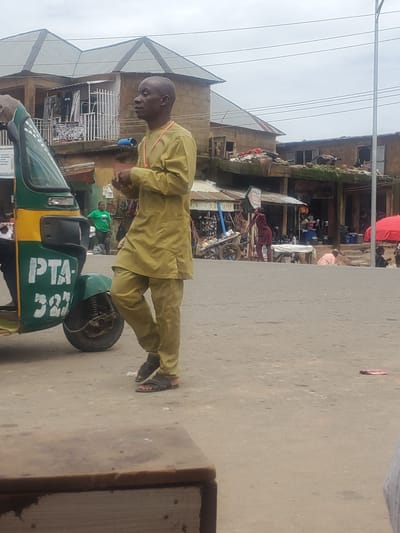 The image depicts a street scene in Gwong, Nigeria. In the foreground, a man in a khaki outfit stands near a green and yellow auto-rickshaw. The auto-rickshaw has "PTA-323" written on its side. Behind the man, buildings with corrugated metal roofs line the street, and several other individuals are visible, some engaged in commercial activities. A woman in a green shirt is standing by the auto-rickshaw, and further in the background, people are gathered around market stalls. A red umbrella is visible to the right, and a street light is present on the far right. The overall environment suggests a bustling urban or semi-urban area with moderate pedestrian and vehicular activity.