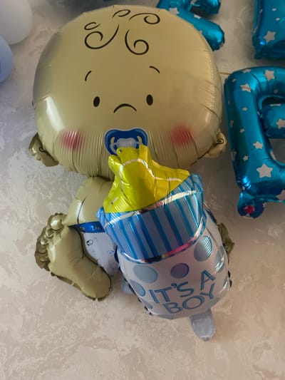 This is a close-up, high-angle shot of several baby-themed balloons against a textured, light-colored wall. The primary focus is a large, tan-colored balloon shaped like a baby's head with black curly hair, a pacifier, and rosy cheeks. To the right of the baby balloon, parts of large blue balloons with white stars are visible, likely forming the number "2" and potentially other numbers. Below the baby balloon, a smaller, blue and white balloon shaped like a baby bottle is prominent, reading "IT'S A BOY" in blue lettering. The balloons appear to be inflated and arranged together, suggesting a celebratory setting such as a baby shower or birth announcement. The location is identified as Istaravshan, Tajikistan.