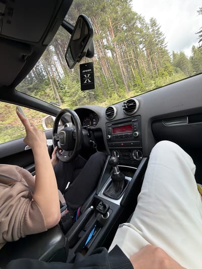 The interior of a car is shown from the driver's perspective, with trees visible through the windows. A person is in the driver's seat, holding a phone in their left hand. Another person, wearing white pants, is in the passenger seat. An "Areon Version" air freshener hangs from the rearview mirror. The radio displays "Audi concert 102.5 MHz." No location information is provided regarding the city of Kasak, Bulgaria.