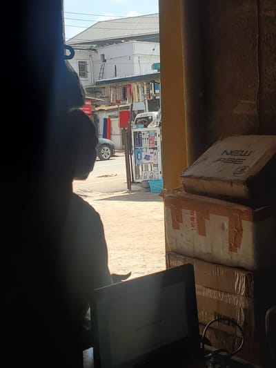 The image depicts a scene observed from inside a shaded structure, looking out onto a sunlit commercial street in Fegge, Nigeria. In the immediate foreground, a person is silhouetted, facing away from the camera and towards a laptop with a glowing screen. The individual appears to have short, dark hair and subtle facial hair visible on the jawline. To the right of the frame, several cardboard boxes are stacked. The top box is light brown with "NEW AGE" printed in dark letters, accompanied by an illegible logo. Lower boxes are taped with brown parcel tape, one showing partial Chinese characters.
Outside the structure, a busy street scene unfolds. The ground is a light-colored, unpaved or dirt surface. A black car is visible on the street. To the left, across the street, a white building with a grey corrugated roof dominates the background. Adjacent to it, another white building features a wooden-framed window and a wooden ladder leaning against its facade. Along the street, market stalls or vendor displays are visible. One partially legible sign reads "RUGS" and "Project & Flowers." A mobile display stand, possibly for electronics, is positioned on the right side of the street, showing various small items and a blue plastic basket at its base. A white van with "UWA ON AIR" text on its side is partially visible behind this display. The overall environment suggests a bustling urban commercial district.