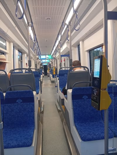 The image shows the interior of a modern tram in Wrocław, Poland. The tram features rows of blue seats with a white wave-like pattern, and grey headrests. The interior is well-lit by overhead fluorescent lights. Metal poles and blue hand straps are visible throughout the tram, indicating it is in motion or prepared for passengers to stand.
In the foreground, on the right side of the tram, a yellow ticket validator machine is mounted on a pole. The machine has a screen displaying information and a slot for tickets. Text on the machine reads "TUTAJ SKASUJ TULET BILET", which translates to "Here validate your ticket".
On the left side of the tram, a person wearing a straw hat is seated, seen from the back. Further down the tram, another person is seated on the right side, also viewed from the back, wearing a light-colored t-shirt. In the distance, more passengers are visible, and the aisle extends towards the front of the tram.
The windows of the tram offer glimpses of greenery outside, suggesting the tram is traveling through a city area with trees. The overall impression is of a clean and functional public transport vehicle.