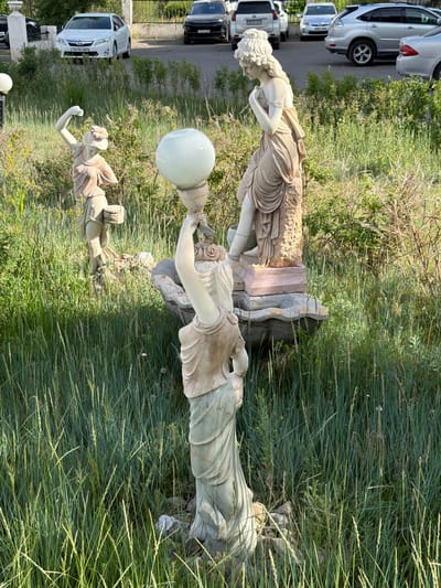 In Ulaanbaatar, Mongolia, an outdoor scene features a garden with several statues of women amidst tall green grass and bushes. In the foreground, a statue of a woman is depicted from the waist up, holding a large, round, translucent sphere on a pedestal. To the left and further back, another statue shows a woman with her arm raised, holding a basket. Behind these statues, a third statue of a woman is visible, with her arms clasped at her chest. Several cars are parked in the background on a paved surface, including a white sedan, a black SUV, and a silver SUV. A metal fence separates the garden from the parking area.
