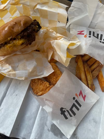 This is a close-up, overhead shot of a fast-food meal placed on a table, as indicated by the wooden texture visible in the top right corner, which is consistent with outdoor seating often found at restaurants in Budapest, Hungary. The meal consists of a cheeseburger in a bun, chicken tenders, and french fries, all served in paper wrappers. The wrappers are white with a checkered pattern in yellow and white for the burger, and plain white with the "fini'z" logo for the tenders and fries. The food is scattered across the table, suggesting it's being consumed or has just been served. The lighting suggests it's daytime, possibly outdoors. No people or actions are visible, only the remnants of a meal.