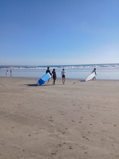 This is a wide-angle, eye-level, outdoor shot of a beach in Kuta, Indonesia. In the foreground, the sand is tan with numerous footprints and small piles of sand, likely from sand crabs. The middle ground shows a calm ocean with small waves breaking and a clear blue sky above. Several people are present on the beach. In the middle distance, two individuals are walking towards the right, each carrying a surfboard. The person on the left is carrying a large blue surfboard, while the person on the right is carrying a white surfboard. To the left of them, two more people are walking on the wet sand closer to the water, with one person appearing to carry a surfboard. Further in the distance, there are other individuals in the water and on the shore. The lighting indicates it is daytime, with the sun casting long shadows on the sand.