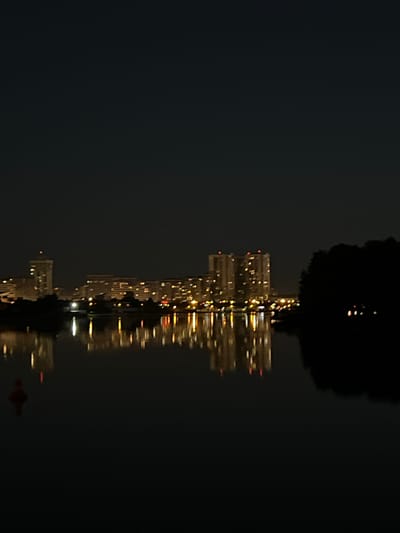 The image shows a nighttime view of a city skyline with buildings and their reflections in the water. The lights from the buildings create a warm glow against the dark sky. The water is calm, reflecting the lights of the city, and a small red buoy is visible in the foreground. The overall scene is serene and captures the beauty of the city at night. The location is a body of water in Minsk, Belarus, likely a river or canal, with apartment buildings visible on the opposite bank.