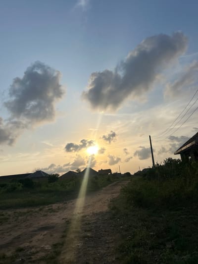 The image depicts a semi-rural or residential area within Debasco, Nigeria, at either sunrise or sunset. The sky displays a gradient of light blue at the zenith transitioning to a warm yellow-orange hue near the horizon, with several scattered cumulus clouds. The sun, a bright luminous disc, is positioned slightly left of center, low on the horizon, emitting distinct rays of light that extend downwards and diagonally to the right.
In the foreground, an unpaved dirt path, approximately 2-3 meters wide, curves gently upwards towards the center-right of the frame. The path's surface appears uneven. Dense green vegetation, composed of various grasses and bushes, flanks both sides of this path, with a more significant concentration along the right edge.
In the mid-ground, several low-rise structures with rounded or sloped roofs are silhouetted against the bright horizon, likely residential buildings. On the far right, a dark utility pole stands vertically, supporting multiple overhead power lines that traverse horizontally across the upper-mid portion of the image. Adjacent to the utility pole, a partial view of a building structure, possibly under construction or in disrepair, is visible on the far right. No individuals, vehicles, or animals are present in the scene.