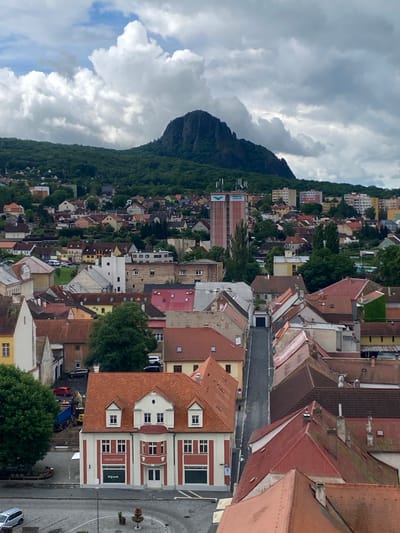 This is a high-angle, exterior, eye-level shot of the city of Bílina, Czechia, on a cloudy day. In the background, a large, dark, rocky mountain dominates the skyline, covered in green trees. Below the mountain, a dense urban landscape of residential and commercial buildings is visible, with varying roof colors and architectural styles. Some buildings are modern, with flat roofs, while others are older, with pitched red roofs. A tall, multi-story building with antennas on top is situated in the middle ground, possibly a telecommunications tower or office building. In the foreground, closer to the viewer, is a street lined with buildings with red tiled roofs. The street appears to be a main thoroughfare, with cars parked along its sides. One building in the foreground, a two-story structure with white and red facade, has a sign that reads "Infocentrum." There are also construction vehicles visible on the left side of the image, including a blue truck and yellow excavator. The overall impression is a mix of urban development and natural landscape, with a sense of everyday life in a European city.