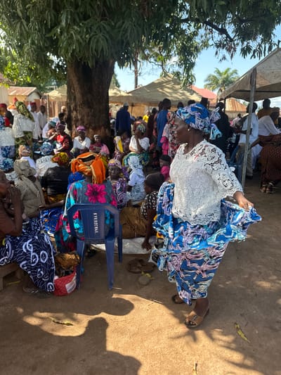 In Mararaba, Nigeria, a group of people are gathered outdoors under a large tree and tents. The ground is dirt. A woman in the foreground is dancing or posing, wearing a white lace top and a blue and purple patterned skirt, with a matching headscarf. She is moving her skirt with her right hand. Many other people, mostly women, are seated on the ground or on chairs, dressed in colorful traditional clothing and headscarves. Some are looking towards the woman in the foreground, while others are engaged in conversation or looking elsewhere. Behind the gathering, there are buildings with corrugated metal roofs and palm trees visible. The scene appears to be a social event or community gathering.