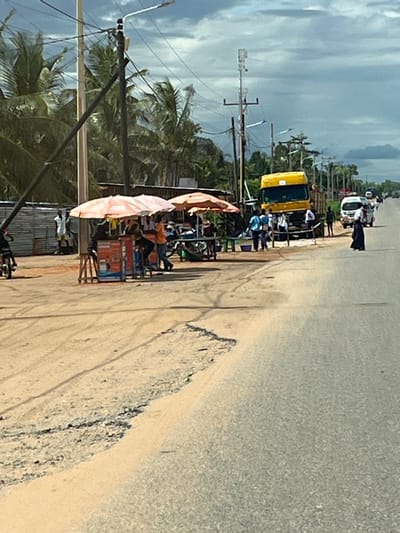 This image captures a roadside scene in Muxara, Mozambique, under an overcast sky. The foreground features a dirt shoulder on the left, displaying tire tracks and merging with a paved asphalt road that extends into the background on the right. An eroded section of the dirt shoulder is visible along the road's edge.

On the left side of the dirt shoulder, two orange and white striped market umbrellas provide shade over impromptu stalls. Under the left umbrella, a small counter displays items in red and blue packaging, with multiple individuals standing around it. One person in an orange shirt appears to be attending the stall. Two wooden stools are positioned nearby. Behind these stalls, several men are visible, and a building constructed from corrugated metal sheets stands. A person on a bicycle is partially visible on the far left, moving away from the viewer.

Further down the road on the right, a yellow truck, identifiable as a semi-trailer cab, is parked partially on the dirt shoulder and partially on the paved road. Several individuals are gathered around the front of this truck. Further along the asphalt road, a woman in a long black garment walks away from the viewer. Additional vehicles, including a white van, are discernible in the distance on the road.

The background is characterized by numerous tall palm trees, indicative of a tropical environment. Multiple utility poles with overhead power lines are distributed along the roadside; one taller pole prominently features cellular antennae.