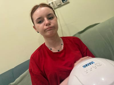 A young Caucasian woman with light skin and reddish-brown hair pulled back is seated, facing the camera with a displeased expression. She is wearing a bright red t-shirt and a silver necklace with four black clover-shaped charms. A white UV nail lamp with a digital display showing "0.00" is visible in the lower right quadrant of the image, with the woman's hand resting near it. The background consists of a pale yellow wall and a light green couch. The overall lighting is soft.