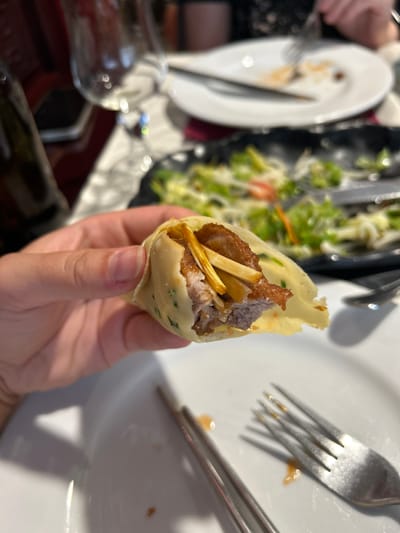 A hand holds a close-up of a food item wrapped in a thin crepe or pancake, with visible fillings of meat and possibly some vegetables or fried noodles. In the background, a white plate with a fork and chopsticks rests on a white tablecloth. Further in the background, a blurry salad is visible on a black plate, and a glass of wine is on the table. The scene appears to be a restaurant setting, possibly in Porto, Portugal.