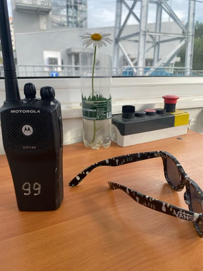 The image depicts an indoor scene, likely on a desk or tabletop, with a wooden surface. In the foreground, a pair of black sunglasses with a pineapple pattern and "VANS" branding on the arm are placed. To the left, a black Motorola CP140 walkie-talkie is positioned, with the number "99" marked on its side. Behind these objects, a clear plastic bottle containing water and a single daisy is visible. Further back, a control panel with several buttons, including a prominent red emergency stop button, is present. The background shows a window with a view of industrial-looking structures and some greenery, suggesting an urban or industrial setting. There is no direct indication of the specific location within Прикамский, Russia, beyond it being an indoor environment with an external view of what appears to be an industrial facility. The scene is static and shows no people or ongoing actions.
