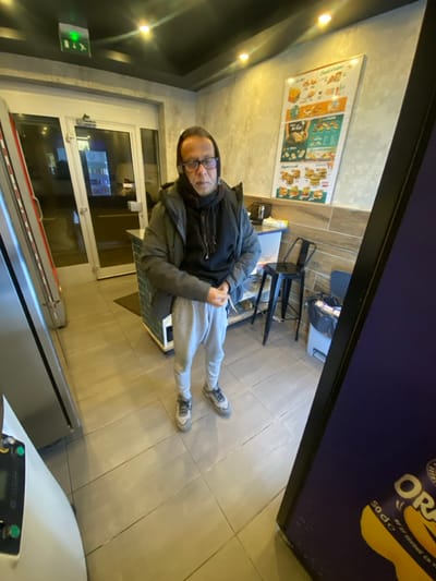 A man wearing glasses, a black hoodie, a gray jacket, and gray sweatpants sits at a table in what appears to be a restaurant. He is looking towards the camera. Behind him are tables and chairs, and a menu board on the wall. A door is on the left side of the image. The shot is taken from a high angle. The frame is partially obstructed by a cooler, obscuring parts of the view.