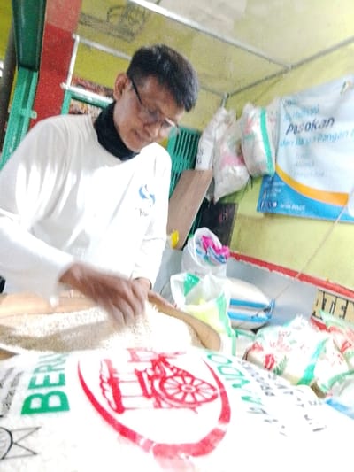 A man wearing glasses and a white long-sleeve shirt, is working in what appears to be a small grocery. He's in front of a bag of rice. He seems to be sorting through a pile of rice, possibly checking the quality or preparing it for sale. Bags, including those containing rice, are stacked behind him, and a sign with food-related text is visible on the wall behind him.