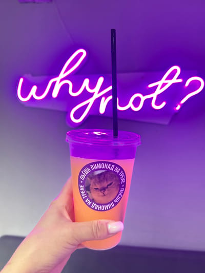 The image shows a hand holding a plastic cup with a straw in front of a purple neon sign. The cup is filled with an orange liquid and has a label with a cat's face and Cyrillic text on it. The text around the cat's face reads "ПЬЕШЬ ЛИМОНАД НА ТРЕНЕ". The hand has light skin and manicured nails. The neon sign is in a script font and seems to say "Why?". The image was taken in Klin, Russia.