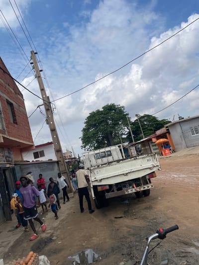 This outdoor street scene in Luanda, Angola, depicts a busy, unpaved road under an overcast sky with patches of blue. The ground is rough, with visible dirt and muddy puddles. The time of day appears to be daytime, possibly mid-morning or afternoon.
In the midground, a white flatbed truck is parked facing away from the viewer, its rear visible. The truck has a license plate that reads "LD 597 HP" and the number "88" marked on its side. To the right of the truck, a portion of a motorcycle handlebar can be seen. Behind the truck, a large tree with dense green foliage stands out, and some small market stalls or kiosks are visible.
Several men are gathered on the street. One man in a light-colored jacket and dark pants is standing near the back of the truck. Closer to the foreground, a group of men in casual attire, including t-shirts, shorts, and one in a purple hoodie, are standing and congregating. Some appear to be looking towards the camera, while others are interacting among themselves. On the far right foreground, what appears to be a basket containing eggs or similar produce is visible.
To the left, a utility pole with multiple power lines extends across the frame, leading towards a red brick building under construction. The scene captures a moment of daily life in a bustling, developing urban area, with people gathered on the street amidst vehicles and local architecture.