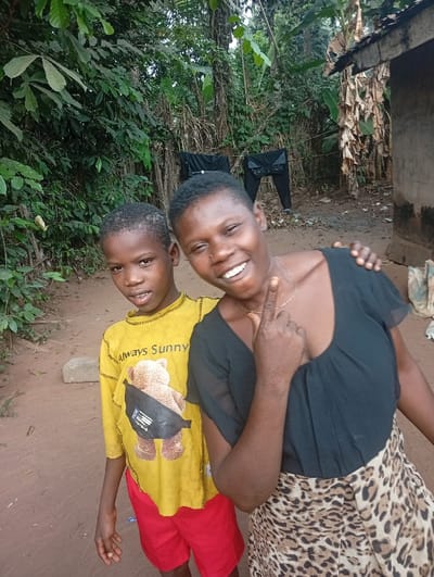 Two individuals are positioned outdoors in Eziudo, Nigeria. On the left stands a male child, wearing a yellow long-sleeved shirt with a brown teddy bear graphic and red shorts. He has short dark hair and a neutral facial expression, looking forward. To his right, an adult female with short dark hair wears a black v-neck top and a leopard-print skirt. She smiles broadly towards the camera, her left index finger raised and bent, and her right hand resting on the child's left shoulder. The ground surface is reddish-brown earth. The background features dense green tropical foliage. To the far right, a rudimentary structure is partially visible, with dried, large plant leaves, consistent with banana leaves, attached or leaning against it. Two pairs of dark trousers are hanging on a visible clothesline in the mid-ground, suspended between trees. A light-colored stone is present on the ground to the left of the child.