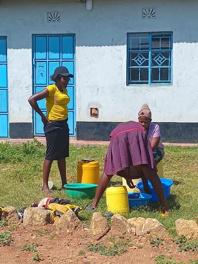 Three women are outside of a building in Malava, Kenya, which is located at latitude 0.53, longitude 34.813. The building is white with blue window frames and a blue door. Two of the women are doing laundry in blue and yellow buckets. One woman is standing in the yard and observing the two others who are kneeling and washing clothes in the buckets. The woman standing is wearing a yellow shirt and a black skirt. The women washing clothes are wearing a burgundy skirt and a headscarf.  There are large rocks around the buckets and other clothes and items on the ground.  The yard is covered in green grass.
