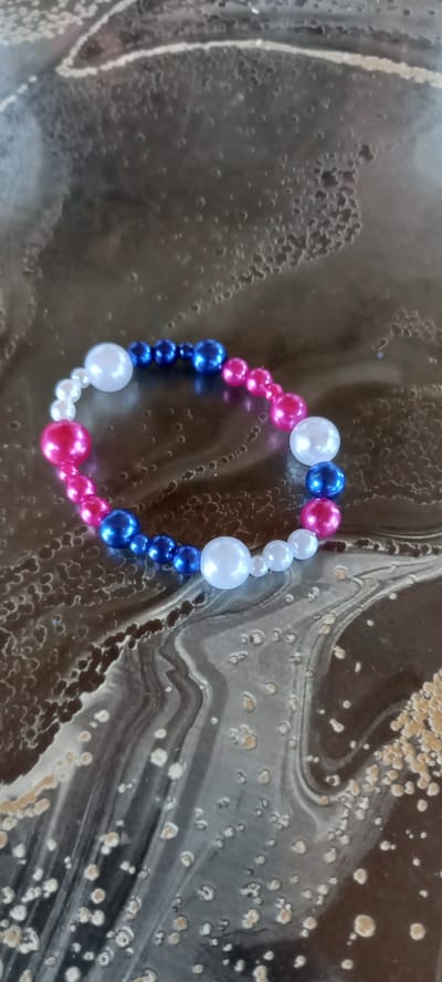 A close-up shot shows a bracelet made of beads of various sizes. The beads are in colors of pink, blue, and white, arranged in a pattern around the bracelet. The bracelet is placed on a dark, textured surface with swirling patterns and small circular indentations. The surface appears to be some kind of resin art or a similar decorative material. The lighting suggests an indoor setting, with no specific time of day or weather cues. There is no visible text in the image.
