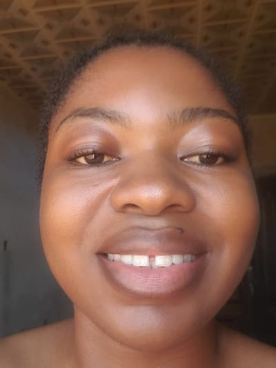 A close-up, front-facing portrait of a young adult female with a rich brown skin tone, captured in Kakau, Nigeria. The subject is smiling broadly, revealing her upper teeth, which show a noticeable diastema between the two central incisors. Her dark, natural curly hair frames her face. She has dark brown eyes with visible dark eyelashes and well-defined, natural eyebrows. A small dark mole is present below her right nostril. Her lips are full and have a natural sheen. The upper background features a textured surface with a repeating geometric pattern, consistent with a decorative ceiling or wall panel, in a warm brown hue. The lower background is dark and indistinct, suggesting an indoor setting with uneven lighting. The subject's bare shoulders are visible in the lower frame.