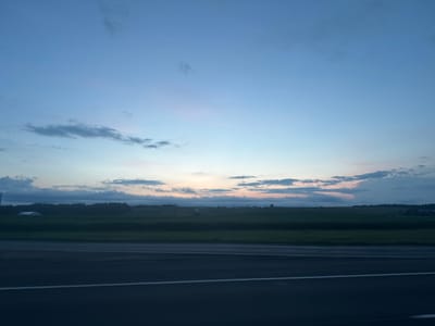 The image captures a twilight scene over a rural landscape near Фаніпальскі сельскі Савет, Belarus. The sky is a gradient of pale blue, transitioning to softer pinks and oranges near the horizon, with scattered clouds. Below the sky, a dark green expanse of fields stretches across the frame, dotted with trees and some distant structures. A white building or structure is visible on the left side, partially obscured by trees. In the foreground, a dark asphalt road with white lane markings runs horizontally, indicating a roadside perspective. The overall impression is one of a serene, rural evening.
