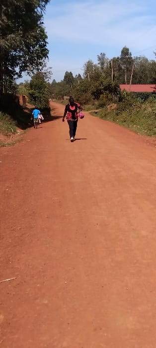 The image shows a dirt road in Bungoma, Kenya, at coordinates 0.562 latitude and 34.803 longitude. A single adult woman in a black shirt and red patterned skirt is walking down the middle of the road away from the camera, carrying a pink bag. A second, smaller figure is walking in the same direction in the distance, in front of the woman. The road is lined with trees and brush on both sides. The sky is blue and there are no clouds.  The scene is rural and the environment is dry.  The road is dusty and the lighting is bright. There are no visible vehicles in the scene.