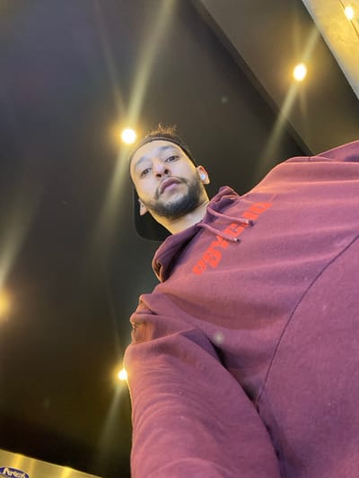 A man looks down at the camera, wearing a maroon hoodie with orange text. He has a dark beard and a light complexion. The background is dark with several bright, circular lights arranged across the top. The camera angle is low, looking up at the man.