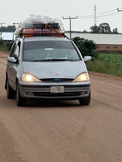 A silver Ford Galaxy minivan is traveling on a dirt road. The minivan's roof rack is loaded with many bottles of drinks, likely soda, covered with white plastic sheeting and secured with straps. The front of the minivan has its headlights on. A license plate on the front of the minivan reads "BSA 198X1 PLATEAU". The environment appears to be rural, with a field of crops on the right side of the road and trees and buildings in the background. There is a power line tower visible in the distance. The sky is overcast.