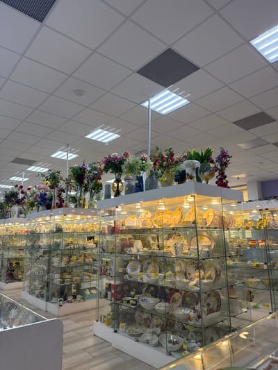 The image depicts the interior of a shop, likely located in Klin, Russia, based on the context. A long row of glass display cabinets stretches into the distance, filled with china, porcelain dishes, cups, and other tableware. The shelves are illuminated with built-in lights. Some shelves are adorned with floral arrangements in vases. The ceiling is a grid pattern of white tiles, with interspersed lighting fixtures and air vents. The floor is light colored. There are no people visible in the image. The shop appears to be specialized in chinaware and related items.