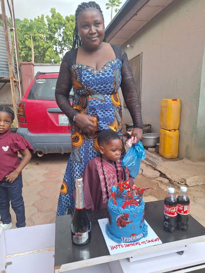 This daylight outdoor photograph, taken in Sabon Tasha, Nigeria, captures a small birthday celebration featuring three individuals.
In the center, a woman with dark skin and braided hair smiles warmly at the camera. She is wearing a blue dress with a vibrant geometric pattern in blue and orange, and black lace long sleeves. She holds a light blue plastic bag containing an indistinct item in her right hand. Standing in front of her is a young boy, likely the birthday celebrant, wearing a maroon patterned shirt over a plain maroon one, looking towards the right. To the lower right of the frame, another young girl with dark skin, dressed in a maroon top with puffy sleeves and blue jeans, gazes directly at the camera.
A blue "Spiderman" themed birthday cake is prominently displayed on a dark-colored table to the right. The cake is decorated with a Spiderman figure and "SPIDERMAN" written on its side. It rests on a white cake board bearing red icing text that reads "HAPPY BIRTHDAY KI. SUCCESS". Beside the cake are two plastic bottles of "Coca-Cola ORIGINAL TASTE" and a bottle of "VELETA" sparkling drink.
The background reveals an outdoor setting. A red car, appearing to be a hatchback or SUV, is parked behind the woman, with "NIGERIA" partially visible on its license plate holder. Beyond the car, lush green trees and foliage are visible under a bright sky. To the left, near a building, are two yellow plastic jerrycans and a metal pot. The ground is a mix of paved and dirt surfaces. The scene conveys a joyful and informal gathering, likely celebrating the young boy's birthday.