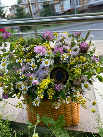 A wicker basket filled with a variety of wild flowers sits on a tiled surface. The basket contains daisies, purple clover, yellow flowers, and a single sunflower. In the background, a silver railing separates the foreground from a blurred image of a building and trees. The overall setting appears to be outdoors, possibly in an urban park or residential area. The image is taken from a slightly elevated angle, looking down into the basket. There are no people or animals visible in the image.
