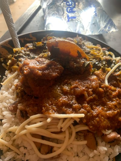 A close-up, overhead shot shows a plate of food, possibly in Shaba, Nigeria. The plate contains white rice topped with a thick, reddish-brown stew. The stew appears to have chunks of meat and leafy green vegetables mixed in. Several pieces of what seem to be pasta or noodles are intertwined with the rice and stew. A silver spoon with an engraved pattern is visible on the left side of the plate, partially submerged in the food. In the background, a plastic water bottle with blue text is reflecting light. The overall scene suggests a meal being served or eaten.