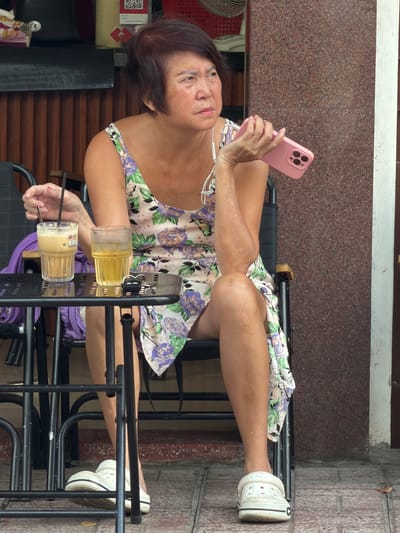 A Vietnamese woman sits at a small black table outside a building with a textured brown facade and wooden slatted siding. She is wearing a light-colored, floral-print dress with purple and green flowers and white clogs. She holds a pink smartphone in her right hand and has earbuds in her ears. On the table are two drinks: one tall glass filled with a light brown iced coffee and another glass with a yellow liquid and ice. A set of keys is also on the table. The background shows a brick-patterned sidewalk and part of a doorway. The scene appears to be a street-level cafe or outdoor seating area in Nha Trang, Vietnam.