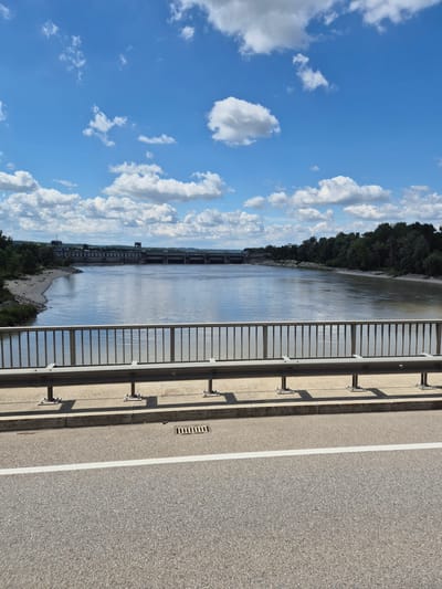 The image is a wide shot of a river with a dam in the distance, taken from a bridge on a sunny day. The sky is blue with scattered white clouds. The river is wide and calm, with a slight brown tint. On the left bank, there is a sandy beach leading to a line of trees. On the right bank, there is a dense forest. The dam is a long, concrete structure spanning the width of the river, with several buildings and sluice gates visible.
In the foreground, the railing of the bridge is visible, with metal posts and a grey metal bar. The road surface of the bridge is grey asphalt with a white line marking the lane. A drainage grate is visible on the side of the bridge.
The image does not depict the city of Braunau am Inn, Austria. The scene appears to be a rural or semi-rural landscape. There are no people visible in the image.