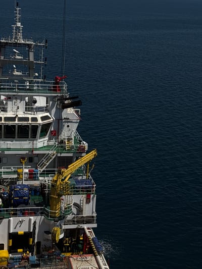 The image shows the side of a large white ship with a green stripe, sailing on a dark blue, rippling body of water. The ship has multiple levels with numerous windows, railings, and various pieces of equipment including antennas, a yellow crane, and what appear to be two large dark-colored exhaust pipes at the top rear of the ship. Several ropes and containers are visible on the deck. The sky is not visible in this shot. The image is taken from an elevated perspective looking down and to the side of the ship.