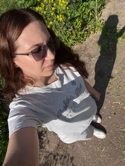 A person lies on the ground outdoors, positioned between vegetation on the left and pavement on the right. The person is oriented with their head towards the lower left and feet towards the upper right corner of the frame. They are wearing sunglasses, a light grey t-shirt, and light grey sweatpants, with white sneakers visible on their feet. The person's brown hair is spread out on the ground, and they are partially lying on yellow flowering plants. The pavement is textured and has scattered leaves on it. A dark shadow, possibly from a tree, is cast across the top portion of the image. The geographical location or city cannot be determined from the information provided in the image.