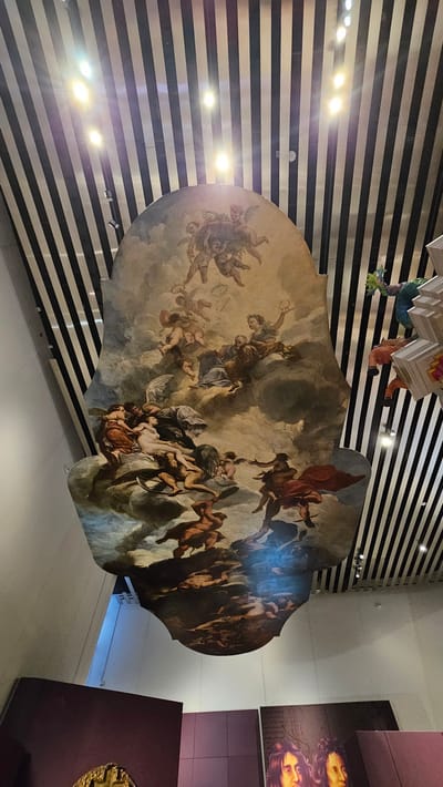 The image shows a ceiling painting in San Bernardino Tlaxcalancingo, Mexico. The painting depicts a classical scene with numerous figures, including winged beings and human forms in varying degrees of dress, rendered in a style reminiscent of Baroque art. The figures are set against a backdrop of swirling clouds and indistinct architectural elements. The painting's shape is irregular, suggesting it might be part of a larger decorative scheme or a unique installation. The surrounding environment includes a black and white horizontally striped wall, a multi-tiered hanging decoration, and a display case.