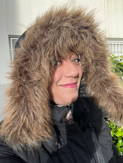 A female subject, appearing middle-aged, is depicted in a close-up, chest-up outdoor portrait, set in Meknes, Morocco. She is wearing a dark (likely black) quilted jacket with a prominent, voluminous, light brown faux fur-trimmed hood that frames her face. Her head is slightly tilted, and she exhibits a closed-mouth smile. Visible facial features include dark eyes with mascara, defined eyebrows, and pink lipstick. A light-colored, possibly patterned, garment is visible beneath the jacket's collar. Strong sunlight illuminates the scene, casting shadows on the subject's right side and jacket. The background features a light-colored architectural structure with visible horizontal window elements on the left, and vibrant green foliage on the right.