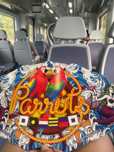 The image shows a close-up view of a circular, colorful graphic that is being held by a person's hand, with the interior of a train car in the background. The graphic features two red macaws facing each other, a ship's wheel, nautical flags, a mermaid, and the text "CARIBBEAN" and "PLAZA INDUSTRIA, 2, SITGES". The train car appears to be moving, with trees and foliage visible through the windows. The seats in the train are grey and blue. The graphic is likely a menu or promotional material for a business located in Sitges, Spain.