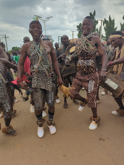 A group of men, identified as "Nzo Cultural Dancers" by the lettering on their attire, are performing in Maigizo, Nigeria. They are dressed in matching leopard-print outfits consisting of shorts and tops, adorned with multiple strands of colorful beads. The dancers are also wearing white sneakers with fringed ankle decorations. Some are playing drums, while others are holding feathered objects. The street scene shows a cloudy sky, utility poles, and other onlookers in the background.