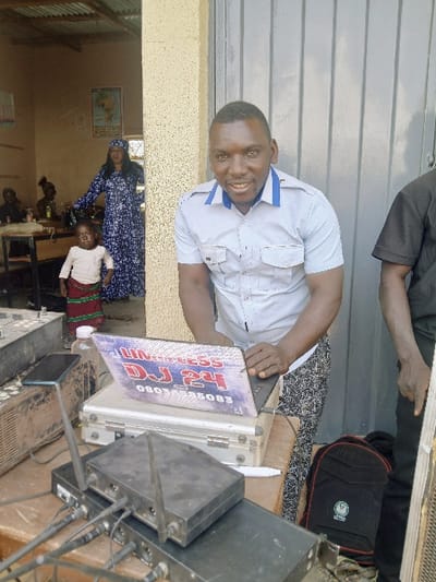 This image captures an outdoor and indoor scene in Masvingo, Zimbabwe. In the foreground, a man with dark skin, wearing a light blue short-sleeved shirt with blue shoulder epaulets and dark patterned pants, smiles directly at the camera. He stands in front of a grey paneled door and operates a silver laptop placed on a wooden table. The laptop screen displays a sticker or graphic with the text "Unipress DJ 24" and "0803445083".

On the table, a black smartphone is propped vertically next to the laptop. Below the laptop is a silver equipment case. In front of this, two black audio receiver units with multiple antennae and connecting cables are visible. A larger piece of dark audio equipment is partially visible to the left. Another individual, partially visible on the far right, wears a black shirt, with a black backpack featuring a green logo resting on the ground nearby.

Behind the main subject, an open doorway reveals the interior of a room with light-colored walls and a corrugated metal ceiling. Inside the doorway, a woman in a long blue and white patterned dress and head covering stands, looking towards the camera. A young child, wearing a light-colored top and a patterned skirt, stands in front of her, also looking towards the camera. Further inside the room, a world map is affixed to a wall, and other individuals are seated at desks or tables.