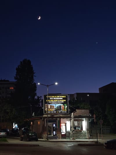 This is a night-time, eye-level, outdoor, full-shot photograph of a street scene in Shumen, Bulgaria. The sky is a deep blue with a crescent moon visible in the upper left quadrant, and a small, bright star or planet in the upper right quadrant. In the mid-ground, a single story building with a stone facade and a brick upper section is illuminated by interior lights. Above this building is a large billboard advertising a real estate company. The billboard text is in Bulgarian and translates to: "QUALITY AND SECURITY WITHOUT PROMISES. WE BUY AND SELL APARTMENTS AND SHOPS. ALL PROPERTIES ARE ACT 16." Below the text are images of apartments and shops, and phone numbers. A street light with two bright bulbs is positioned in front of the billboard. To the left of the building, a tall, dark tree silhouette is visible against the night sky. Several cars are parked on the street in front of the building and in the background. The street surface appears to be asphalt with some reflections from the lights. The scene conveys a sense of urban life after dark.