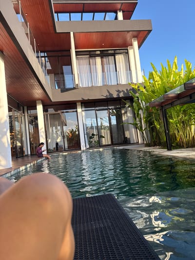 The image depicts a modern villa with a swimming pool, bathed in the warm light of a setting sun. The villa features a multi-level structure with dark gray accents, rich brown wooden ceilings, and extensive glass windows. Lush green plants, likely tropical in nature, flank the poolside, creating a vibrant contrast with the architecture. The swimming pool, with its clear, dark water, reflects the surrounding environment and the sky. In the foreground, the edge of a black lounge chair is visible, and a person, a young woman wearing a purple and black swimsuit, is seated at the edge of the pool with her feet in the water, looking at her phone. Another person's leg, in the immediate foreground, is out of focus, suggesting the photo was taken from a reclining position on the lounge chair. The scene suggests a tranquil and luxurious vacation setting, likely in a warm climate. While the image is not specifically identifiable as being in Kediri, Indonesia, the architectural style and the presence of tropical vegetation are consistent with a tropical Asian setting.