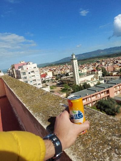 This elevated photograph presents a panoramic view of Meknes, Morocco, under a clear blue sky with scattered white cumulus clouds. In the immediate foreground, a human arm, dressed in a yellow sleeve and wearing a black wristwatch with a silver-colored buckle, holds an open orange-colored aluminum beverage can with Arabic script (identifiable as "Glaam" and "برتقال" (orange)). The hand rests on a textured concrete parapet exhibiting widespread algae and lichen growth. A white cigarette butt is positioned on the parapet to the left of the can. The midground reveals a dense urban environment characterized by numerous multi-story buildings, predominantly residential, in various shades of white, beige, pink, and with red-tiled roofs. A prominent, tall, light-colored mosque minaret with a green-tiled spire rises distinctly above the surrounding structures. Adjacent to the minaret, a large, multi-story building complex with brown facades and flat roofs is visible. The background features a range of green, undulating hills or mountains extending towards the distant horizon.