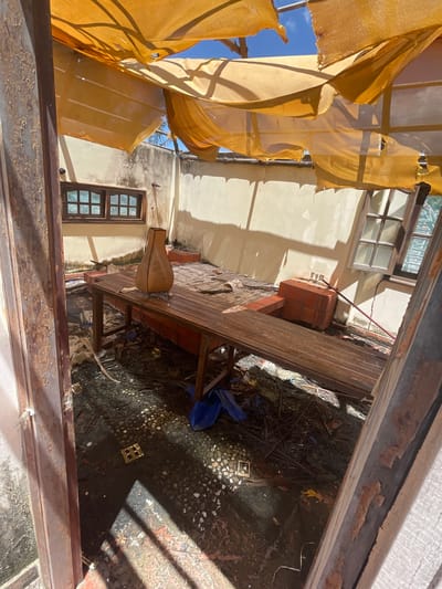 In Phu Thuy Ward, Vietnam, a partially destroyed open-air structure is depicted. The ceiling is composed of torn, yellow fabric draped over a metal frame, with large sections ripped open, revealing a blue sky with clouds. The walls are a light beige, with some areas showing signs of water damage and mold. Two windows with wooden frames and multiple panes are visible on the back wall.

In the center of the room, a long, wooden table sits on a platform. On the table is a tan, vase-shaped object. The floor is cluttered with debris, including plastic sheeting, leaves, and small, light-colored objects. Two terracotta brick structures are positioned against the back wall, one to the right of the table and another smaller one to the left.

The foreground shows damaged, peeling paint on what appears to be the frame of an entranceway. The overall scene suggests the aftermath of a storm or significant damage.