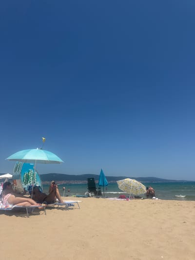 The image depicts a sunny day at a beach, likely in Nesebar, Bulgaria, given the context. The scene is dominated by the vast expanse of a clear blue sky on the left and the sandy beach on the right. The ocean is visible in the background, with gentle waves and a few people swimming. In the foreground, beachgoers are relaxing under umbrellas.

There are several beach chairs occupied by people. One person is lying on a chair under a blue and white striped umbrella with "Sprite" branding visible. Another person is seated on a chair near the water, with a yellow flag attached to a pole nearby. A yellow and white patterned umbrella with lemon designs is also visible, along with another blue umbrella. The overall atmosphere suggests a leisurely summer day at the beach. The lighting indicates it's daytime, and the clear sky suggests good weather. The presence of umbrellas and beach chairs confirms the setting as a popular tourist destination. Text visible includes "Sprite" and "свежо" (fresh in Bulgarian).